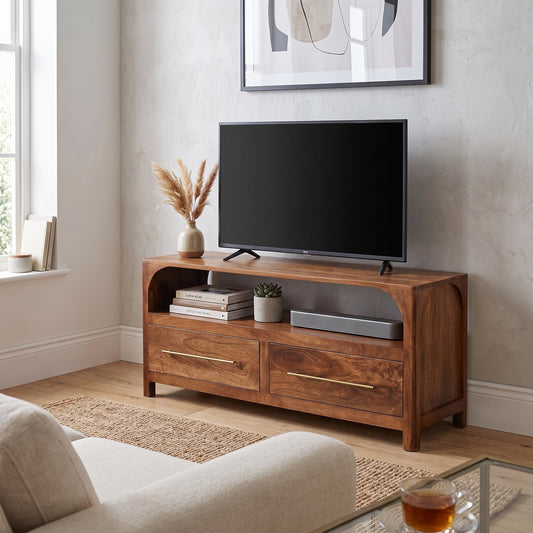 A medium brown wooden TV stand with two drawers and open shelving, styled in a living room setting with a black flat-screen TV, books, and a small potted plant.