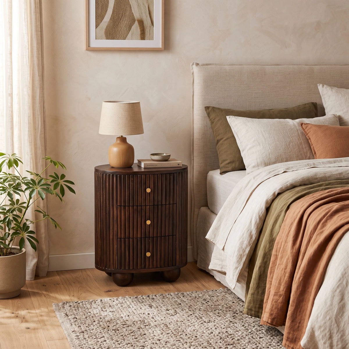 A fluted 3-drawer curved bedside table in solid mango wood with a velvet mocha finish, styled in a warm bedroom setting next to a bed with a small lamp on top.