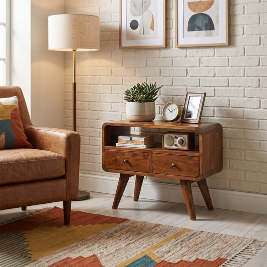 A mid-century modern wooden sideboard with two drawers and an open shelf, styled in a living room next to an armchair and a brick wall.