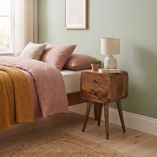 A rustic wooden nightstand with two drawers and tapered legs stands beside a bed in a styled bedroom. The nightstand holds a small potted succulent, a book, and a white ceramic table lamp with a beige shade.