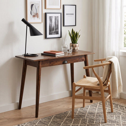 A mid-century modern wooden writing desk with a single drawer, styled in a bright home office next to a wooden chair with a cream throw blanket.