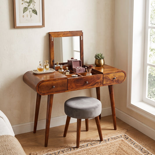 A wooden vanity table with an attached mirror, three drawers, and open top storage, styled with cosmetics and accessories. A round gray upholstered stool sits in front.