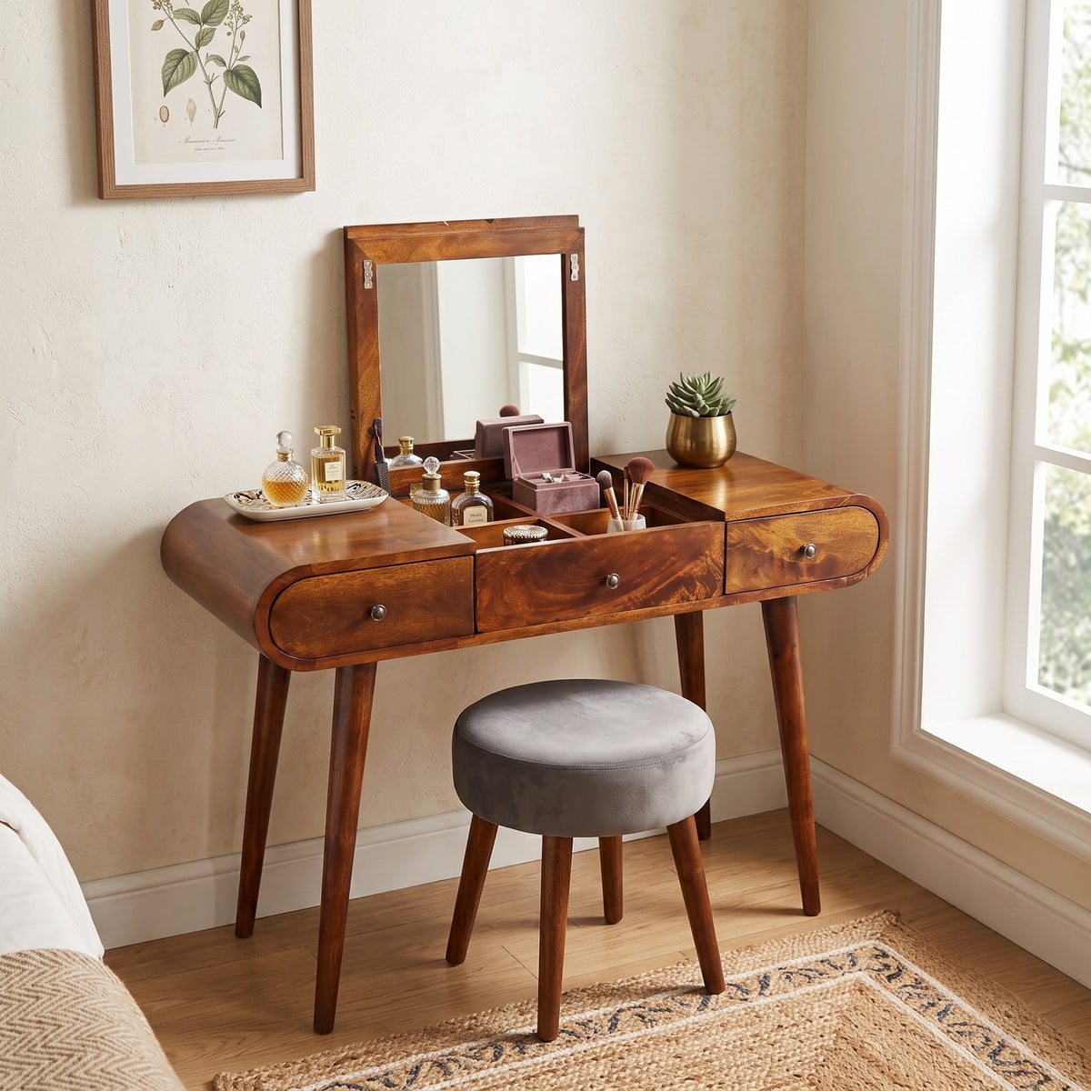 A wooden vanity table with an attached mirror, three drawers, and open top storage, styled with cosmetics and accessories. A round gray upholstered stool sits in front.