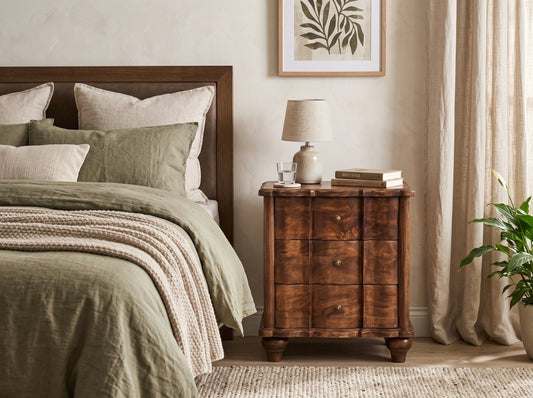 A solid mango wood 3-drawer bedside table in a roasted almond finish, styled in a relaxing bedroom next to a bed with olive green linens.