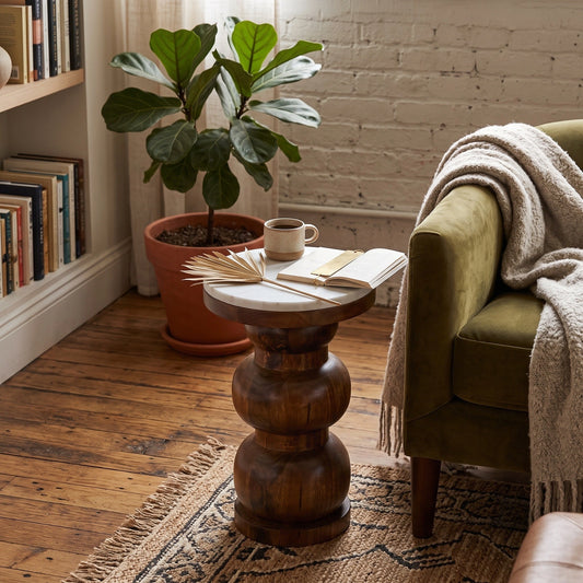 A rustic round wooden, marble top side table with a carved pedestal base and a light natural stone top, styled in a cozy room next to an olive green armchair with a cream throw blanket.