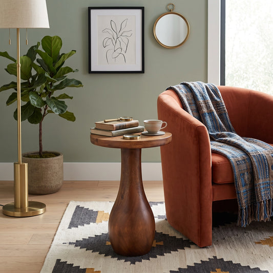 A round wooden side table with a sculptural pedestal base in a styled living room setting next to an orange armchair with a blue plaid throw blanket.