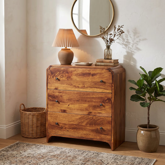 A three-drawer wooden chest of drawers with a warm medium-brown finish, styled in a cozy room corner with a table lamp, books, a mirror, a potted plant, and a wicker basket.