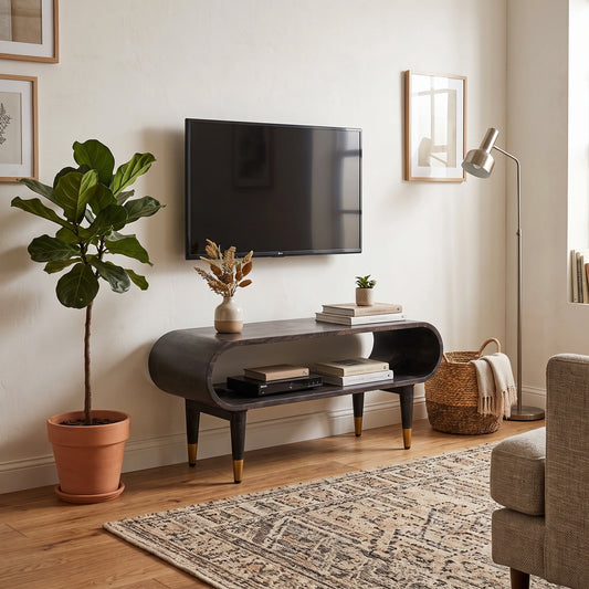 A modern curved media console in a dark wood finish with gold-tone legs, styled in a living room setting with a wall-mounted TV, a potted fiddle-leaf fig, a floor lamp, and a patterned rug.
