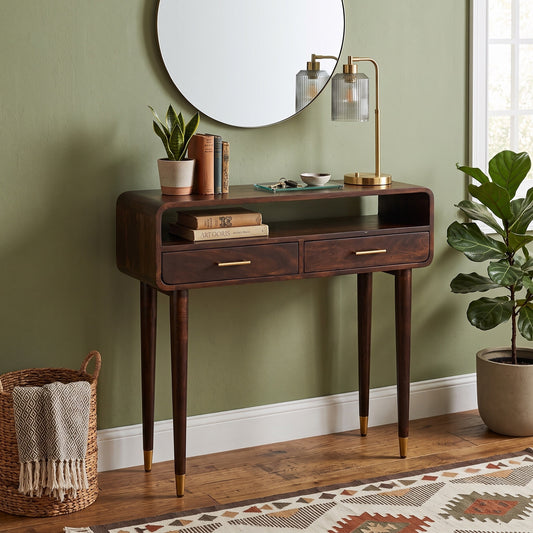 A mid-century modern dark wood console table with two drawers and open shelving, styled in a living room setting with a round mirror, brass desk lamp, books, and potted plants.