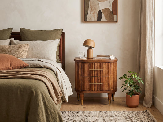 A solid mango wood ribbed bedside table in an amber oak finish, styled in a warm bedroom setting next to a bed with a small dome lamp on top.