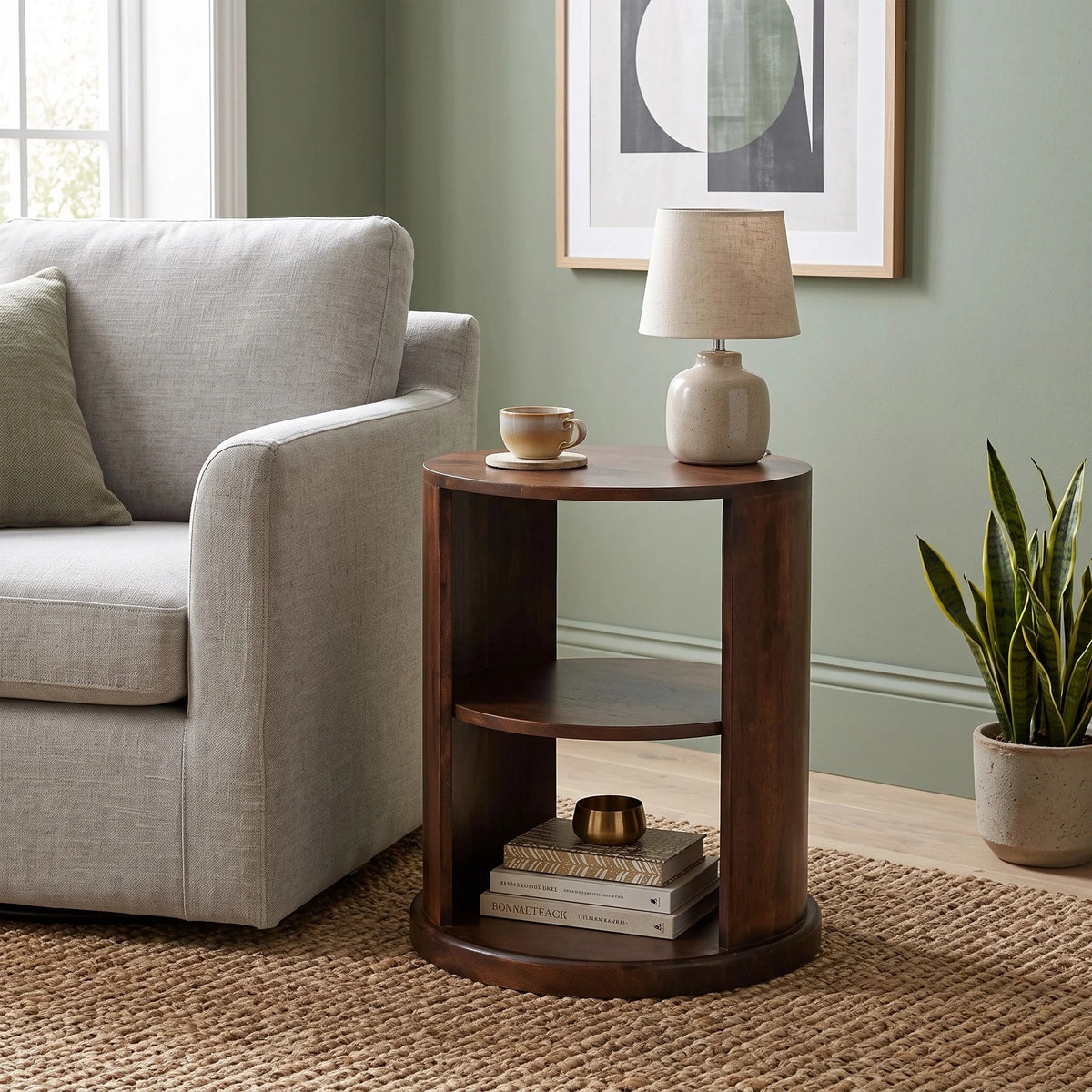 A round dark brown wooden end table with two shelves, styled in a living room next to a light grey armchair. The table holds a small ceramic lamp and a cup, with books on the bottom shelf.
