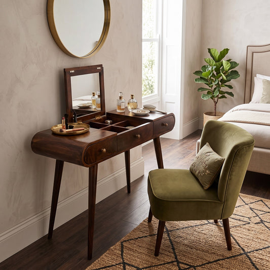 A dark wood vanity table with an attached mirror and storage compartments, styled in a bedroom next to an olive green velvet accent chair.
