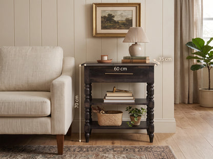 A solid mango wood 1-drawer turned-leg side table in a dark carbon ash finish, styled in a living room next to a light beige sofa with a lamp and decor on top. White dimension lines and text indicate the table's height as 70 cm, width as 60 cm, and the lamp top depth as 35 cm.