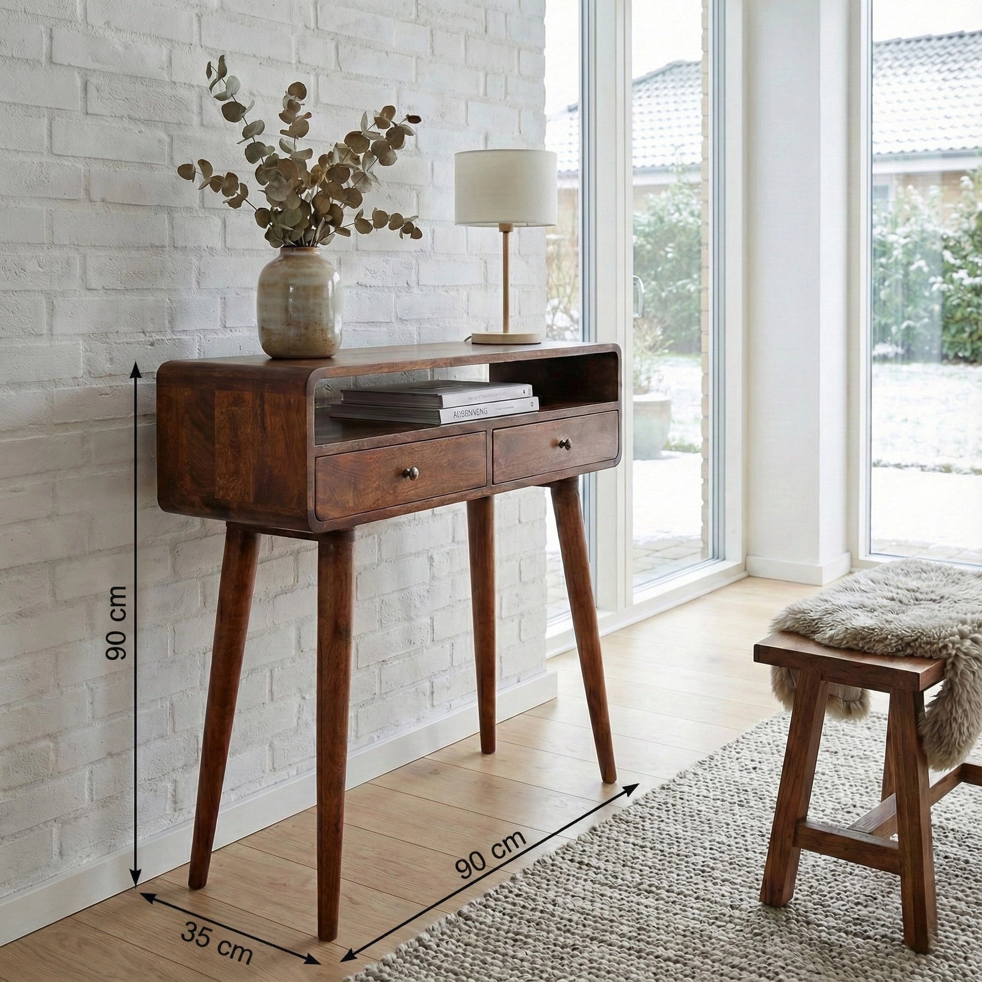 A mid-century modern wooden console table with two drawers and tapered legs, shown in a bright room with white brick walls, dimension lines indicating 90 cm height, 60 cm width, and 35 cm leg width, a vase with eucalyptus, and a lamp on top.