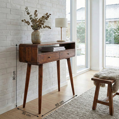 A mid-century modern wooden console table with two drawers and tapered legs, shown in a bright room with white brick walls, dimension lines indicating 90 cm height, 60 cm width, and 35 cm leg width, a vase with eucalyptus, and a lamp on top.