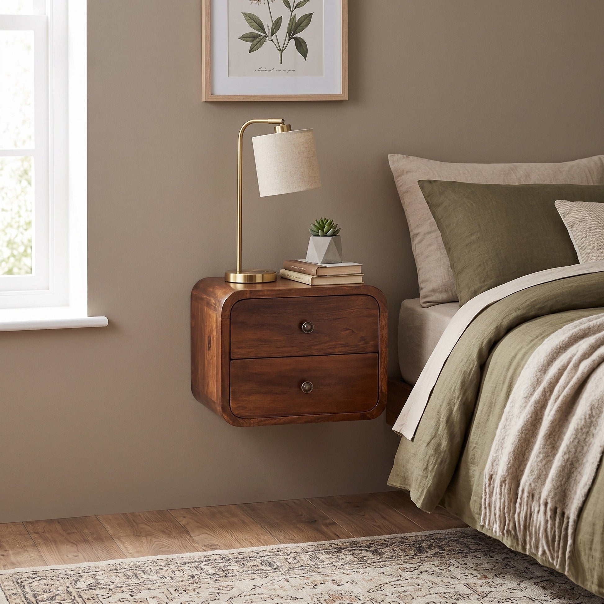 A floating wooden nightstand with two drawers in a warm medium brown finish, styled in a bedroom next to a bed with olive green bedding and a brass table lamp with a white shade.