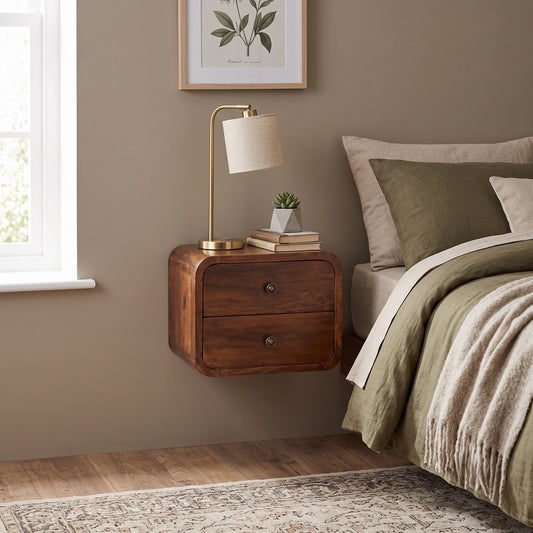 A floating wooden nightstand with two drawers in a warm medium brown finish, styled in a bedroom next to a bed with olive green bedding and a brass table lamp with a white shade.