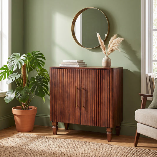 A mid-century modern wooden credenza or entryway cabinet with vertical slats and two brass handles, styled in a room with a green wall, a potted monstera plant, a round mirror, and a chair.