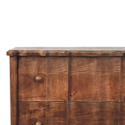 Close-up of a panelled solid mango wood chest in roasted almond, highlighting the scalloped top edge, the wavy vertical paneling on the drawers, and a metal knob.