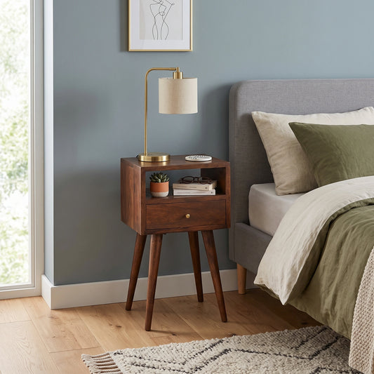 A mid-century modern wooden nightstand with a single drawer and open side shelf, styled next to a bed in a bedroom with a blue-gray wall, brass lamp, and wood flooring.