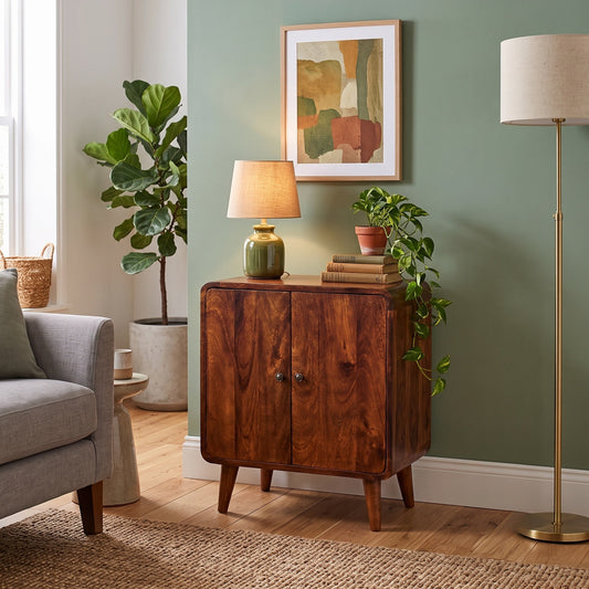 A mid-century modern wooden side cabinet in a warm medium brown finish, styled in a living room setting with a table lamp, books, a potted plant, and a floor lamp against a sage green wall.