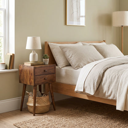 A warm wood bed frame with beige linens and pillows, styled in a cozy bedroom next to a matching wooden nightstand with a white lamp and small plant.