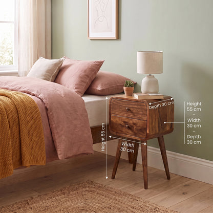 A wooden bedside table with two drawers and tapered legs is shown next to a bed in a styled bedroom. White dimension arrows and text label the table's height, width, and depth.