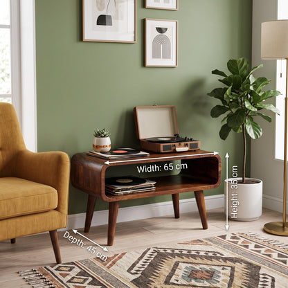 A wooden media unit placed in a styled living room corner, with overlaid dimension labels showing width 65 cm, height 85 cm, and depth 45 cm. The room includes a mustard chair, potted plant, floor lamp, patterned rug, and green wall.