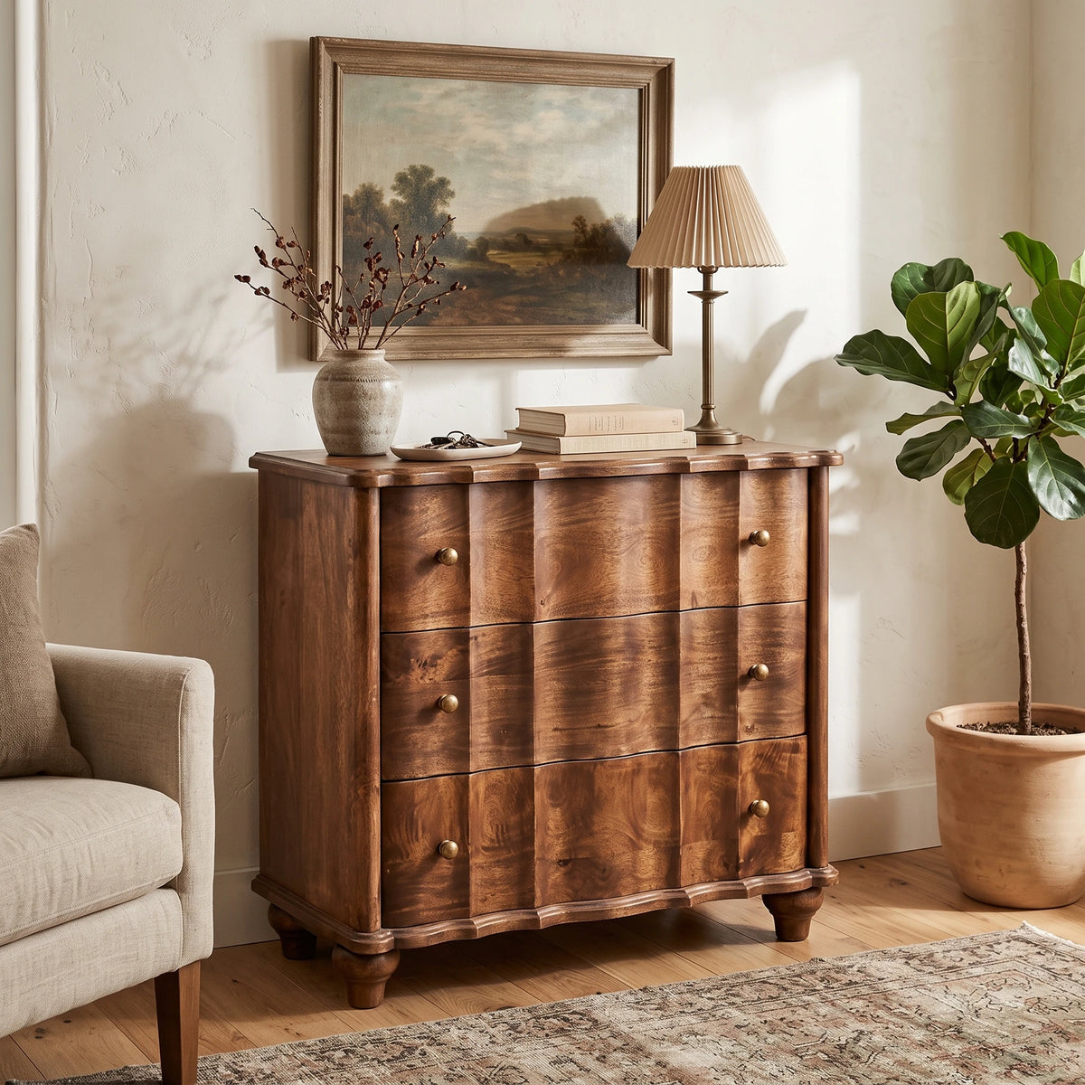 A panelled solid mango wood chest in a roasted almond finish, styled in a warm room setting alongside a framed landscape painting, a table lamp, and a potted fig tree.