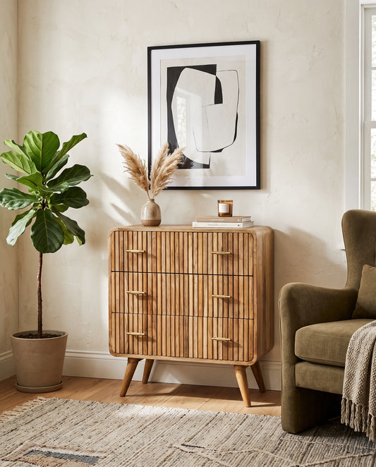 A ridged solid mango wood chest of drawers in a natural oak finish, styled in a cozy room setting with a potted fiddle leaf fig, an armchair, and framed art.
