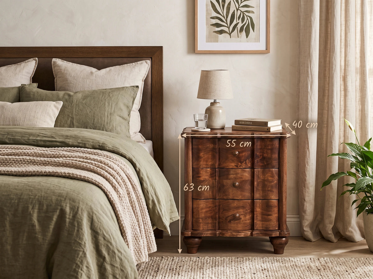 A solid mango wood 3-drawer bedside table in a roasted almond finish, styled in a relaxing bedroom next to a bed with olive green linens, with text overlays indicating dimensions of 55 cm width, 40 cm depth, and 63 cm height.