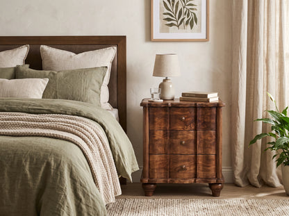 A solid mango wood 3-drawer bedside table in a roasted almond finish, styled in a relaxing bedroom next to a bed with olive green linens.