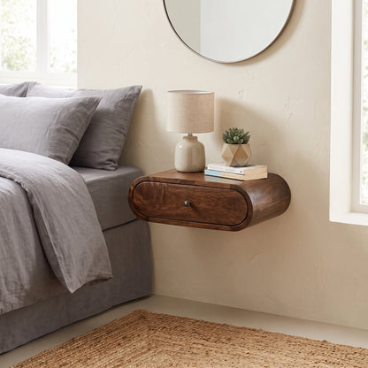 A floating dark wood nightstand with a single drawer, mounted on a beige wall in a bedroom next to a bed with gray bedding. The nightstand holds a beige table lamp, a small potted succulent, and a stack of books.