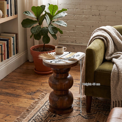 A round wooden, marble top end table with a carved pedestal base, shown in a cozy living room setting next to an olive green armchair. The table has dimension overlays indicating a width of 27 cm, a depth of 27 cm, and a height of 45 cm.