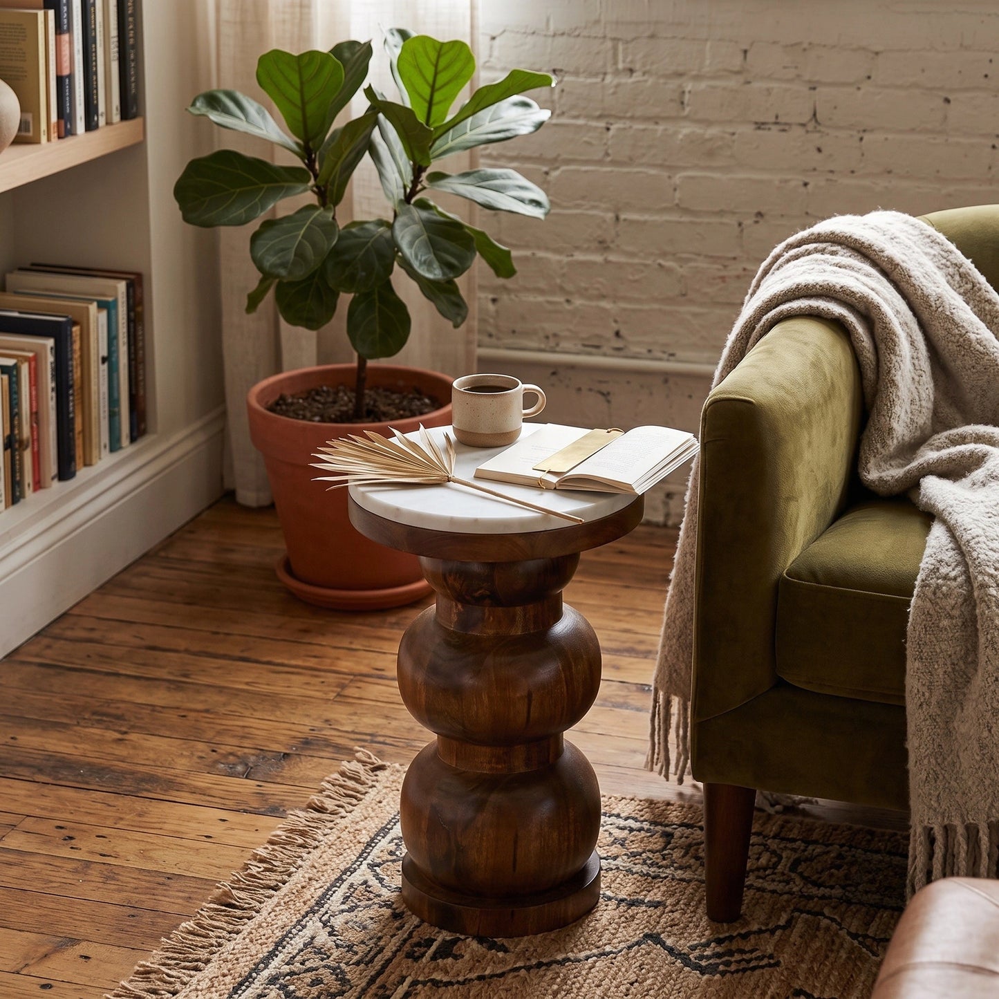 A rustic round wooden, marble top side table with a carved pedestal base and a light natural stone top, styled in a cozy room next to an olive green armchair with a cream throw blanket.
