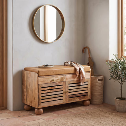 A rustic wooden shoe rack with a tan leather seat cushion, styled in a warm, neutral-toned room with a round gold-framed mirror, a potted plant, and a woven basket.