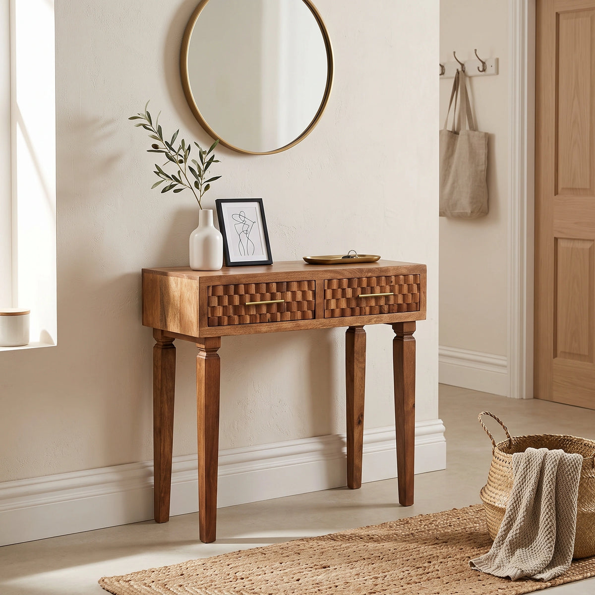 A wooden console table with two drawers and turned legs, styled in a warm, neutral-toned entryway with a round mirror, vase, framed art, and woven decor.