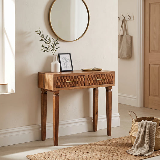 A wooden console table with two drawers and turned legs, styled in a warm, neutral-toned entryway with a round mirror, vase, framed art, and woven decor.