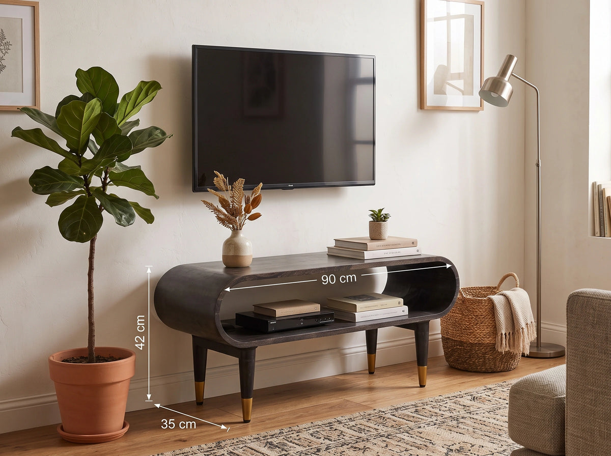 A dark gray curved media console with open shelving and tapered gold-tone legs, shown in a styled living room setting with a TV above, plant, lamp, and dimension callouts indicating 90 cm width, 42 cm height, and 35 cm depth.