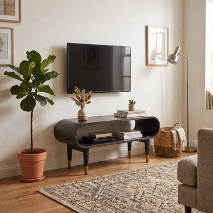 A modern curved media console in a dark wood finish with gold-tone legs, styled in a living room setting with a wall-mounted TV, a potted fiddle-leaf fig, a floor lamp, and a patterned rug.