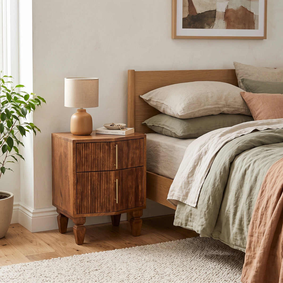 A warm medium-brown wooden bedside table with two drawers and brass handles, styled next to a bed with neutral linens in a softly lit bedroom.