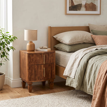 A warm medium-brown wooden bedside table with two drawers and brass handles, styled next to a bed with neutral linens in a softly lit bedroom.