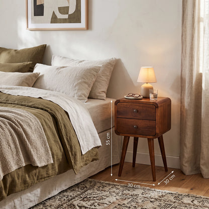 A wooden two-drawer nightstand beside a bed in a styled bedroom, with overlaid dimension lines showing 55 cm height and 30 cm width/depth. No person or animal is present, but the real bedroom environment is visible, so this is Lifestyle rather than Product/Packaging.