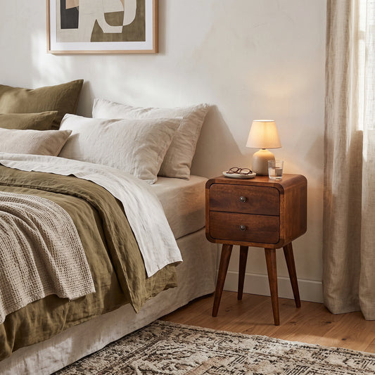 A styled bedroom scene featuring a mid-century modern wooden nightstand with two drawers and tapered legs, styled next to a bed with neutral-toned linens and a lit table lamp.