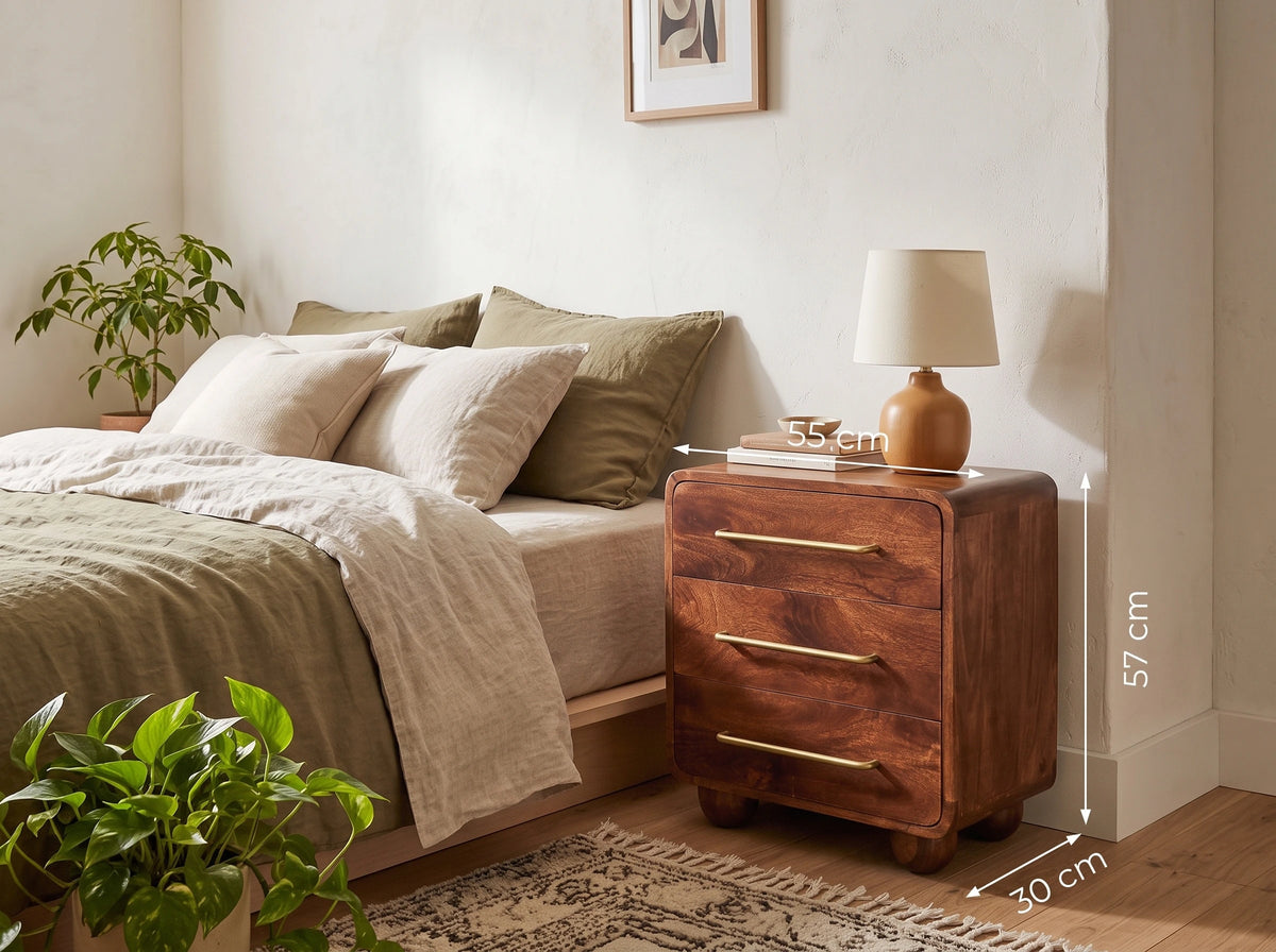 A wooden three-drawer chest with measurements in a warm medium brown finish stands beside a bed in a styled bedroom. The nightstand has brass-tone handles and a small table lamp on top.