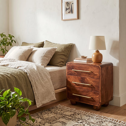 A wooden three-drawer chest in a warm medium brown finish stands beside a bed in a styled bedroom. The nightstand has brass-tone handles and a small table lamp on top.