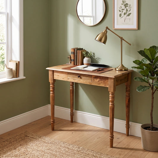 A rustic wooden writing desk with a single drawer and turned legs, styled in a home office setting with a brass desk lamp, books, and plants against a sage green wall.