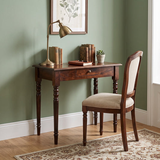 A dark brown wooden writing desk with a single drawer and turned legs, paired with a matching upholstered wooden chair in a styled room with sage green walls, a brass desk lamp, and books.